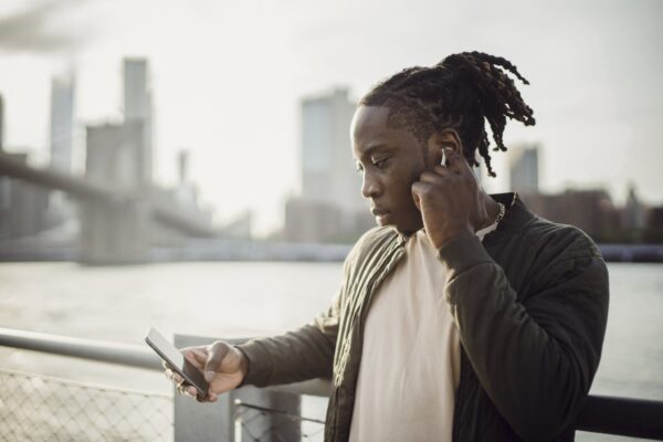 Thoughtful adult African American guy in wireless earphones listening to music while surfing internet on cellphone and standing leaned on hand near fence on bridge over city river