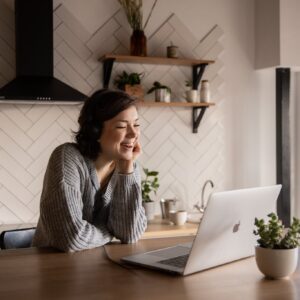 Young cheerful female smiling and talking via laptop while sitting at wooden table in cozy kitchen