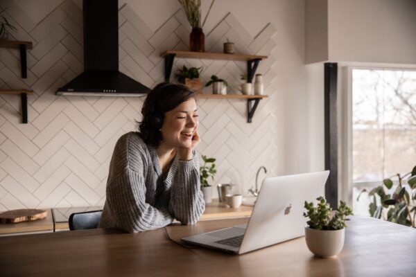 Young cheerful female smiling and talking via laptop while sitting at wooden table in cozy kitchen